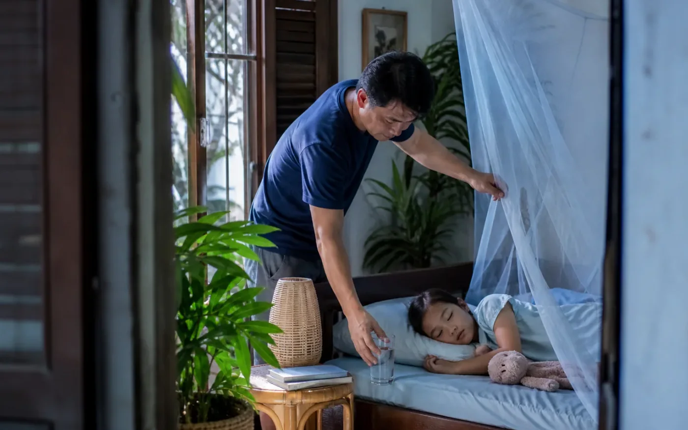 Một người cha Việt Nam đặt nước cạnh con đang nghỉ dưới màn chống muỗi