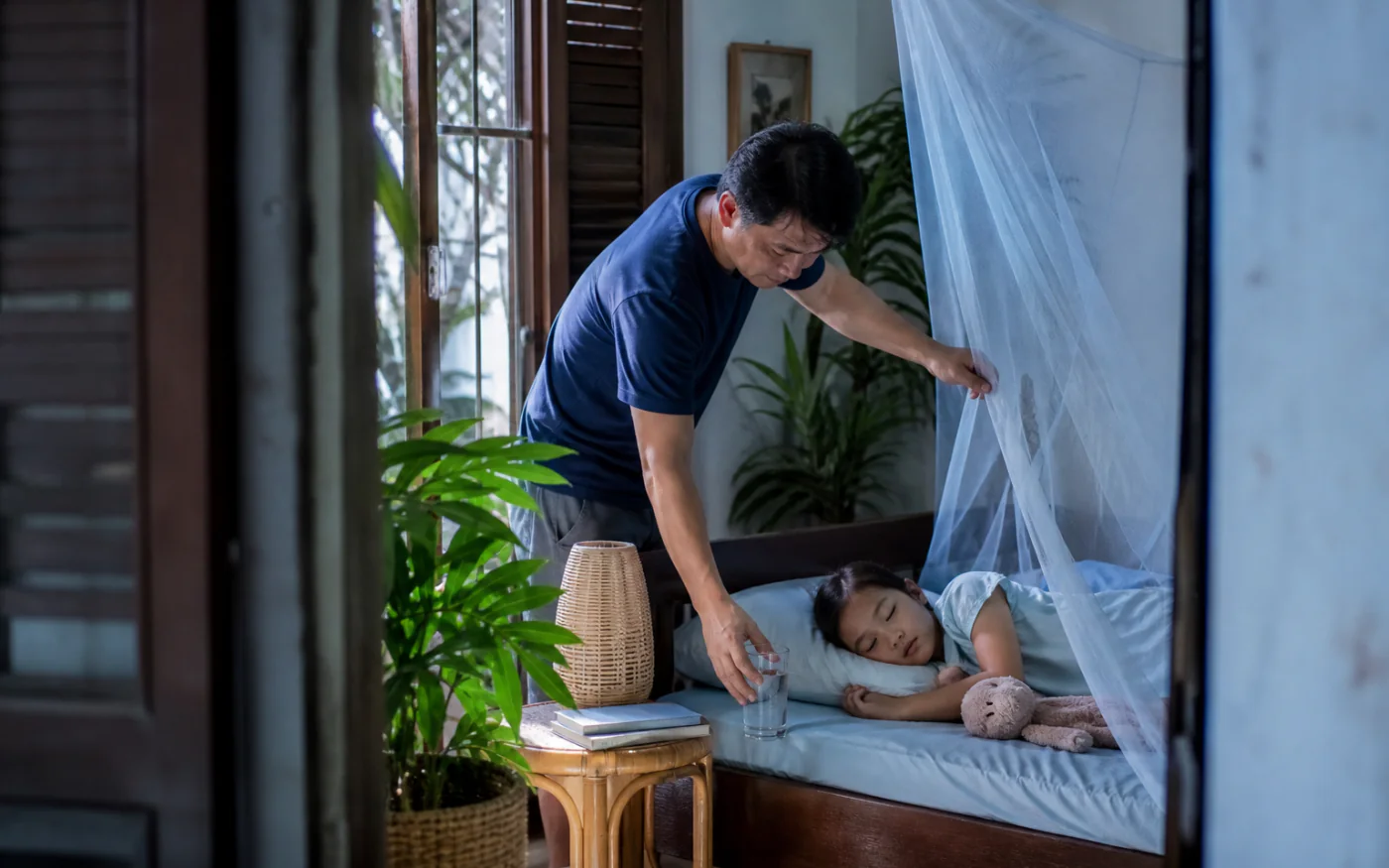 Một người cha Việt Nam đặt nước cạnh con đang nghỉ dưới màn chống muỗi